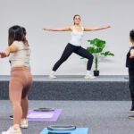 Group fitness class following an instructor on a small stage, practicing standing poses on exercise mats.