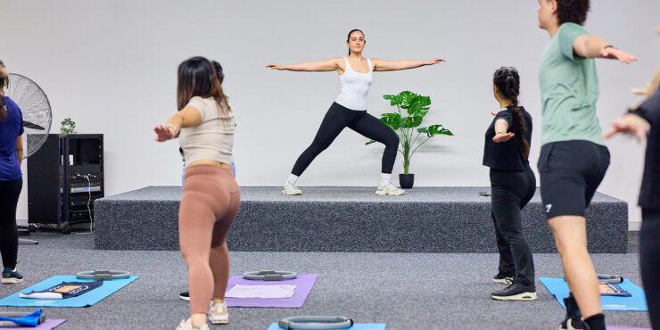 Group fitness class following an instructor on a small stage, practicing standing poses on exercise mats.