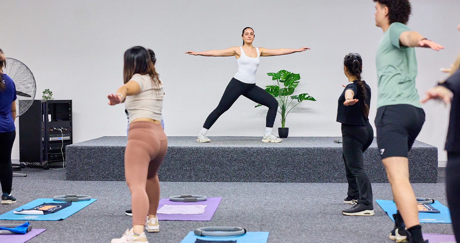 Group fitness class following an instructor on a small stage, practicing standing poses on exercise mats.