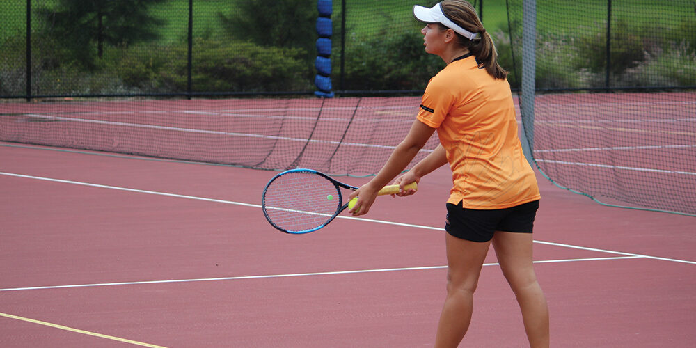 Student wearing an orange t-shirt holds a tennis racquet