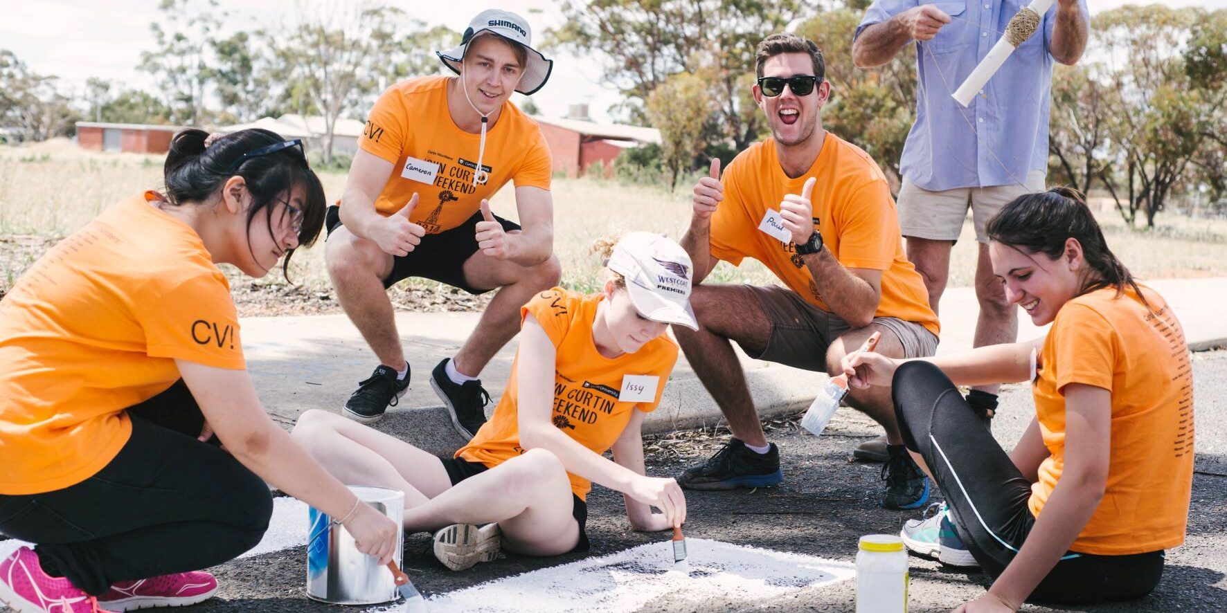 Curtin Volunteer students painting on the ground and smiling. Some students have their thumbs up