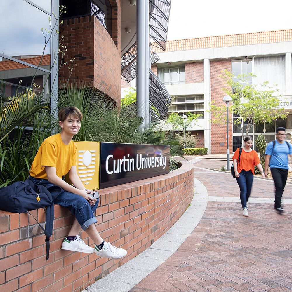 Man sitting on wall with Curtin University sign, with two people walking in the background