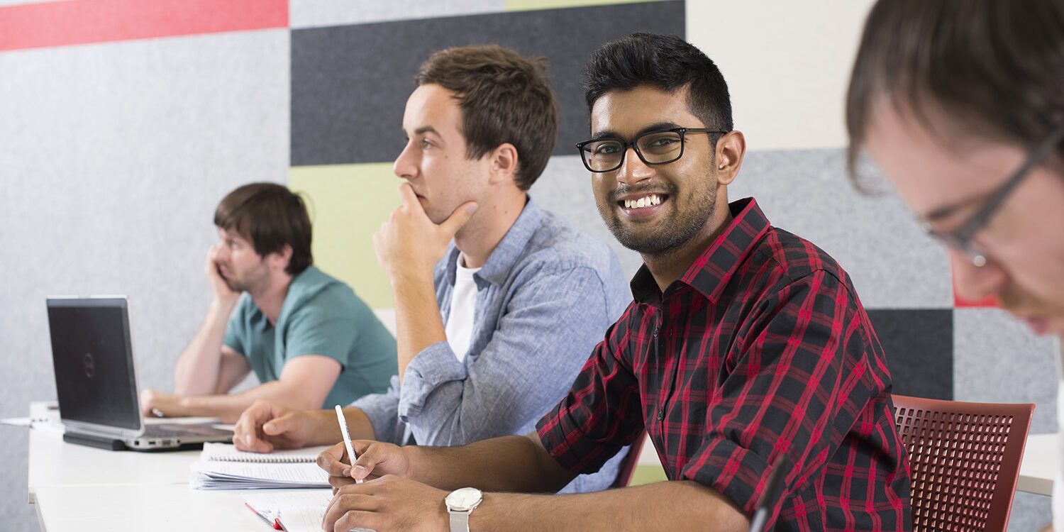 male student sitting amongst other students who are doing work in class. Male student is the only one smiling at the camera