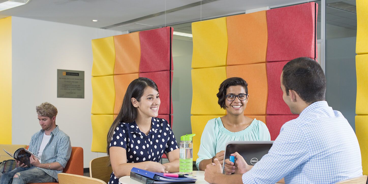 Three students studying at table with man reading in background