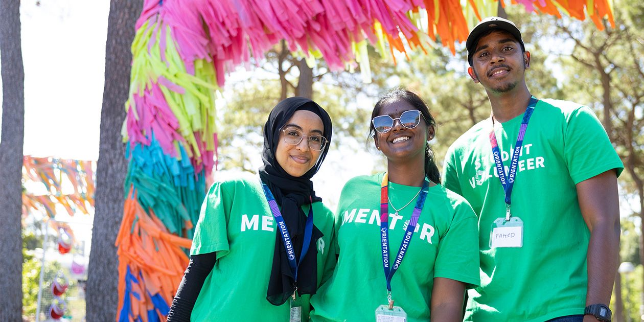 A group of three students wearing green New to Curtin Mentor tshirts and smiling while standing in front of a colourful arbor.