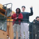 Group of people standing on a large yellow industrial vehicle, waving.