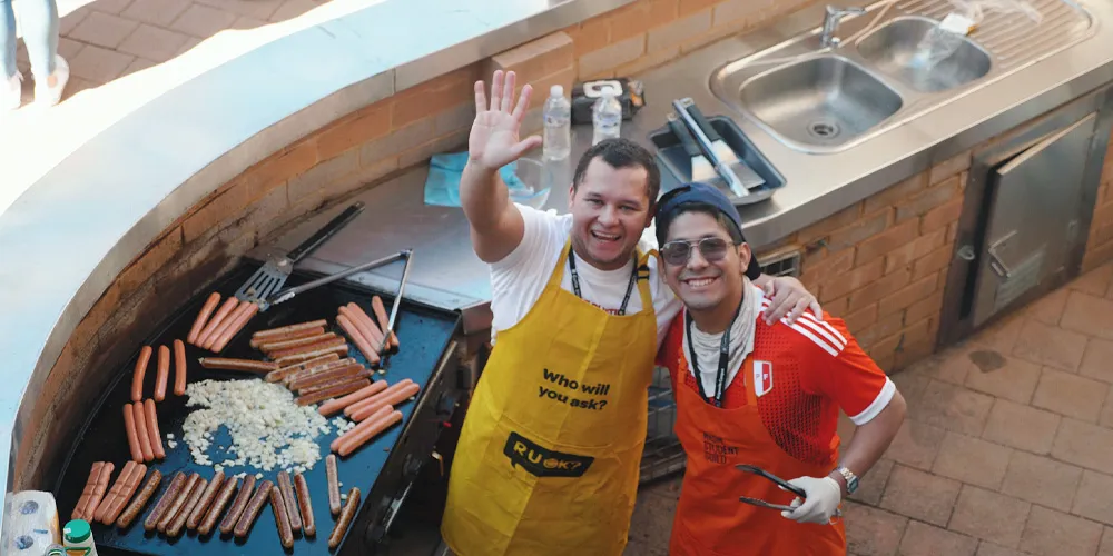 Cooking station with two people grilling sausages and onions on a barbecue. - play video