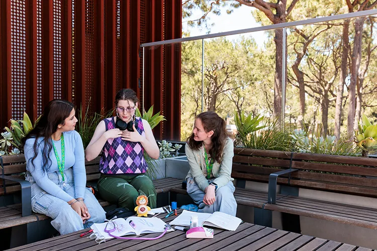 Three people sitting together on outdoor benches, talking and looking at items laid out for study or discussion.