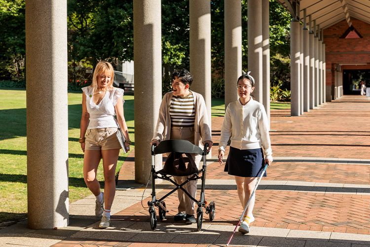 Three people walking together along a covered outdoor walkway, including one using a walker and another using a white cane.
