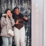 Two students posing with a phone in front of a photo booth backdrop.