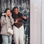 Two students posing with a phone in front of a photo booth backdrop.