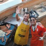 Two people standing at an outdoor grill, holding tongs and waving, with sausages cooking on the barbecue.