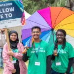 Student volunteers holding an Orientation sign and rainbow umbrella outdoors at a campus event.