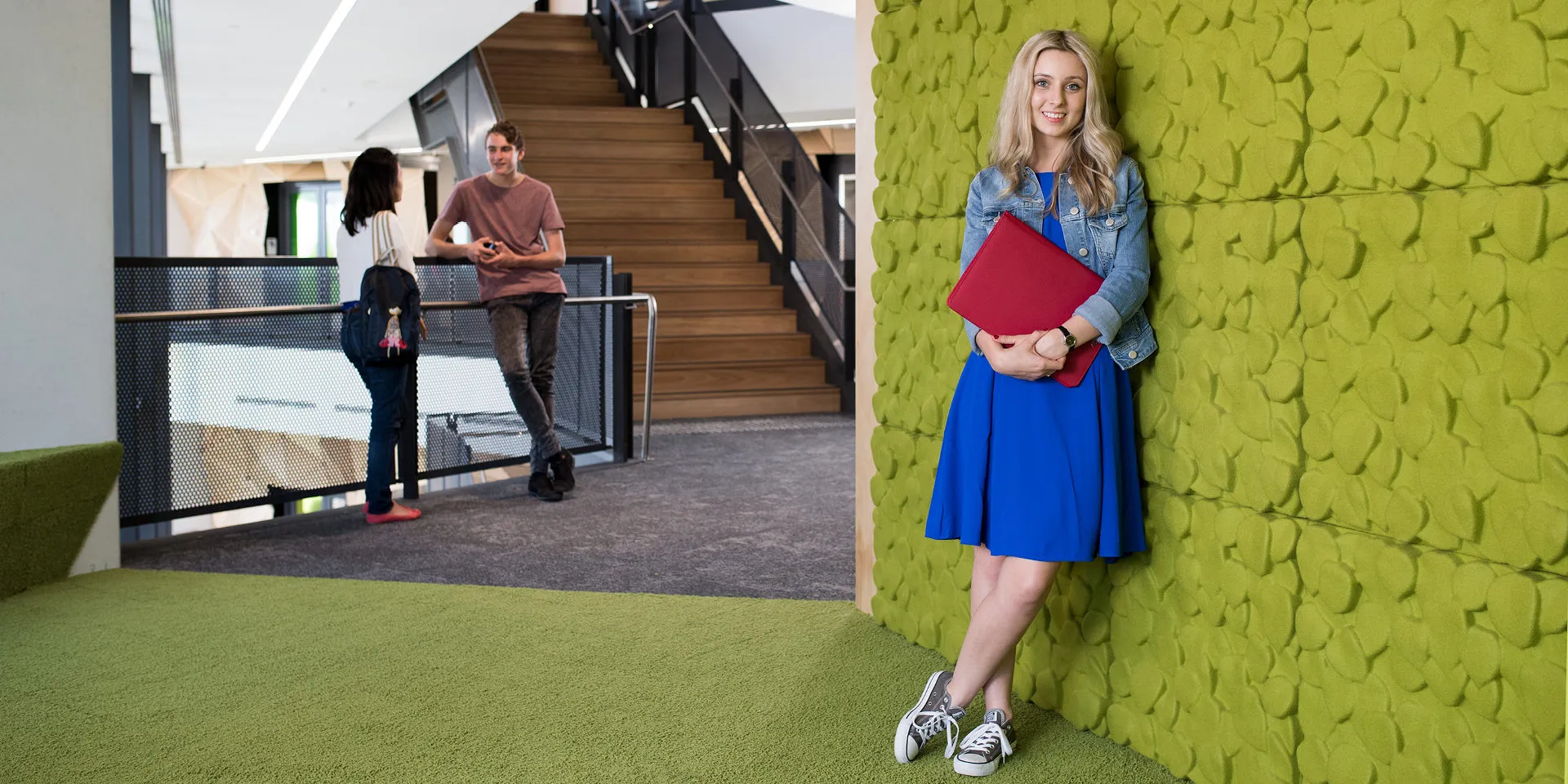 A young female student, leaning against a green wall, holding a red file. Looking at the camera and smiling.