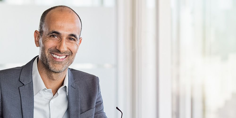 Happy mature business man holding spectacles in modern office. Successful senior businessman in formals sitting in meeting room with phone and tablet. Smiling man in suit in a elegant office.