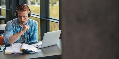 A male in a denim shirt with headphones on, sat at his desk with his laptop