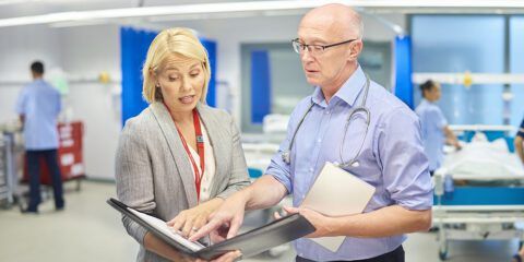a female businesswoman chats to a senior doctor on a busy hospital ward