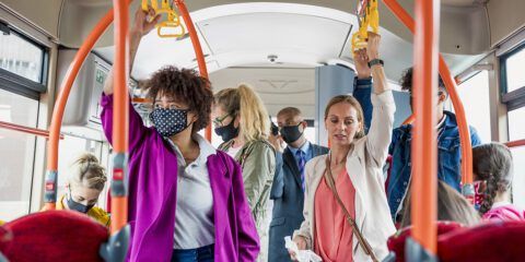 Mixed age and ethnicity passengers on a bus wearing protective masks during the Covid 19 pandemic. One woman is not wearing a mask and is holding a tissue.