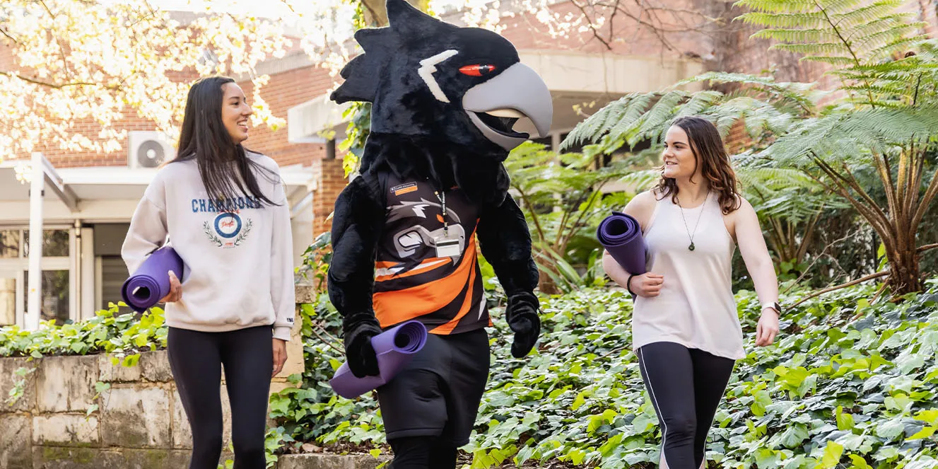 Two students carrying yoga matts walking on campus with the Curtin Carnaby's mascot