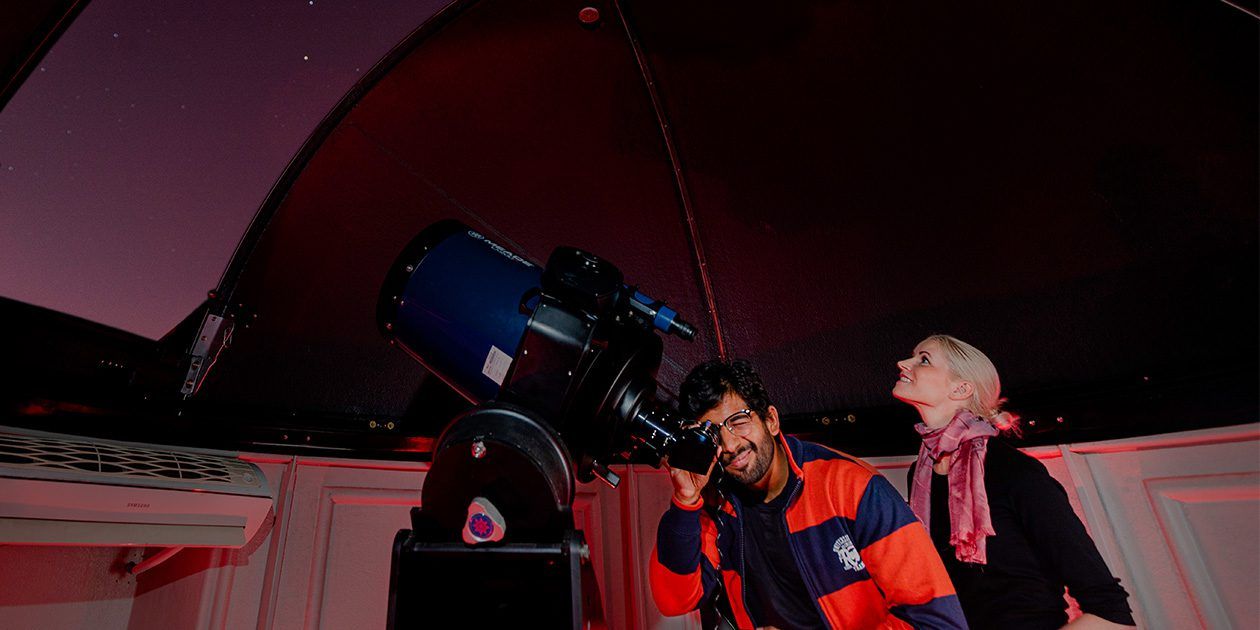 Male and female student looking through a telescope from an observatory at night