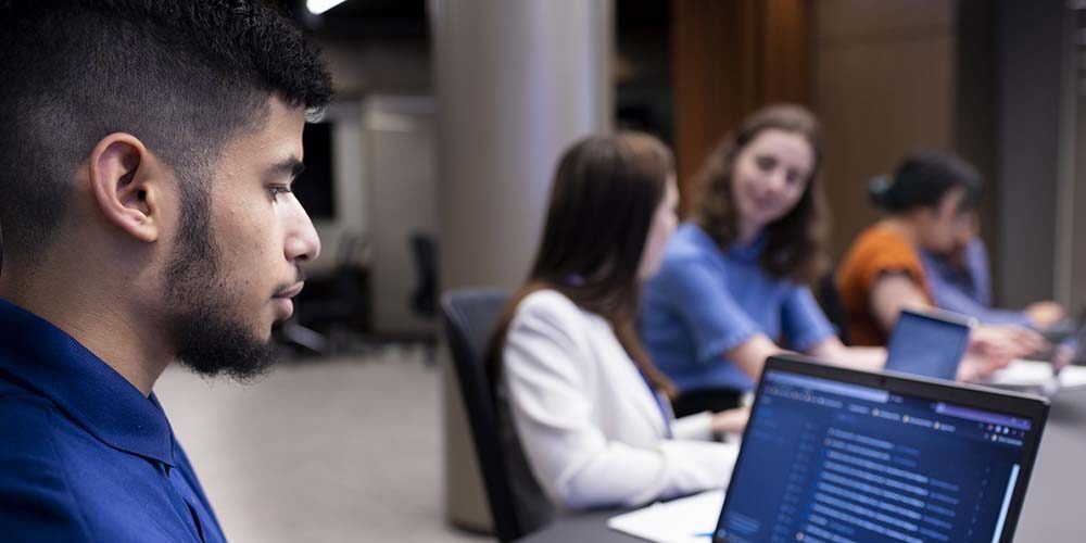 Male student at a laptop with two female students in the background.