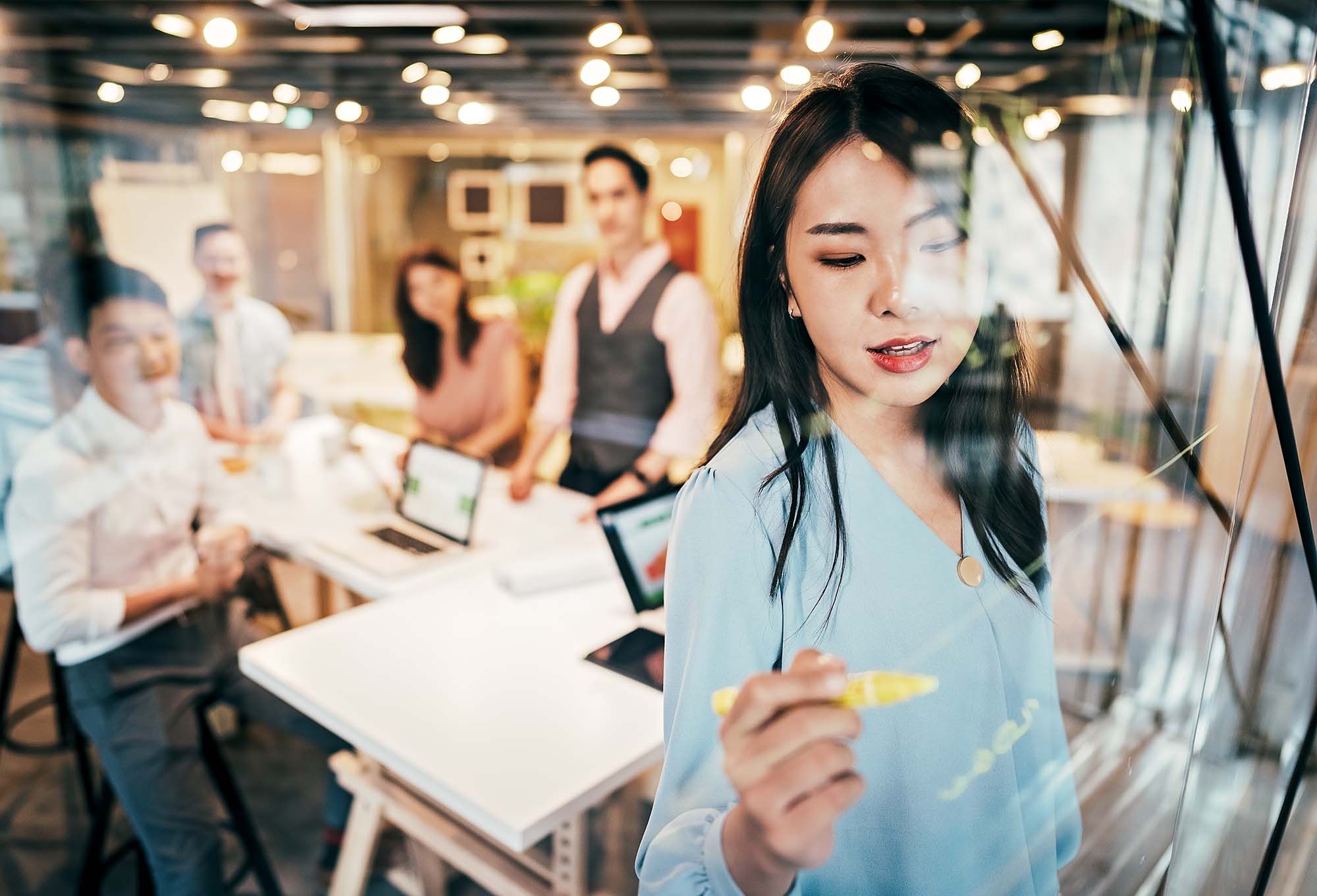 Female in a blue shirt taking meeting notes on an office window, with colleagues in the background.