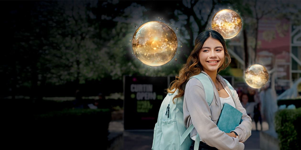 Young woman with long hair, wearing a backpack, holding a book, stands smiling in an outdoor setting.