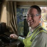 A man with light skin and short dark hair in a work vest and safety glasses drives a train.