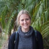 curtin student Emily Cousin standing against a backdrop of palm leaves and dressed in a dark coloured top and jacket