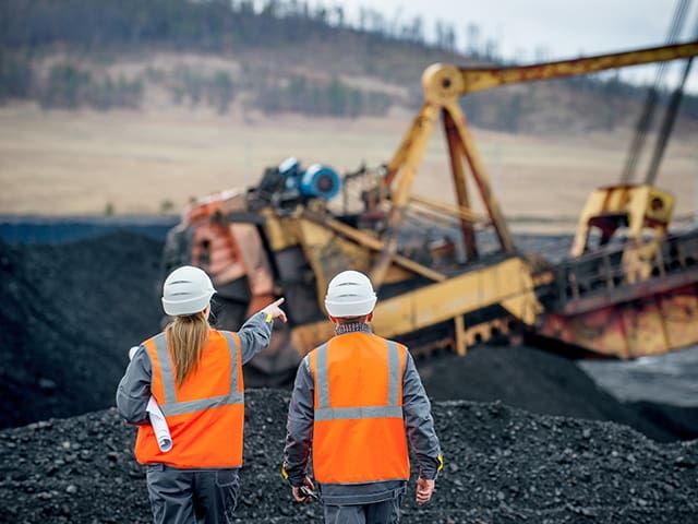 Two workers wearing safety gear, standing on a coal site, with one pointing towards large mining machinery in the background.