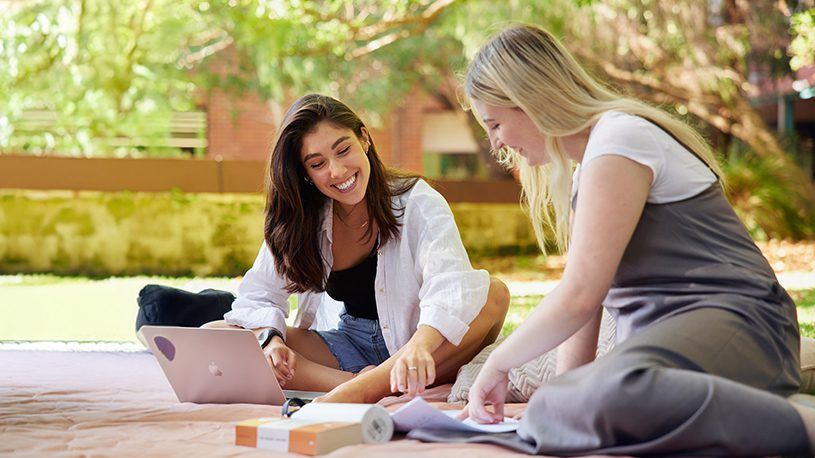 two female students sitting on a picnic blanket in the sun, studying