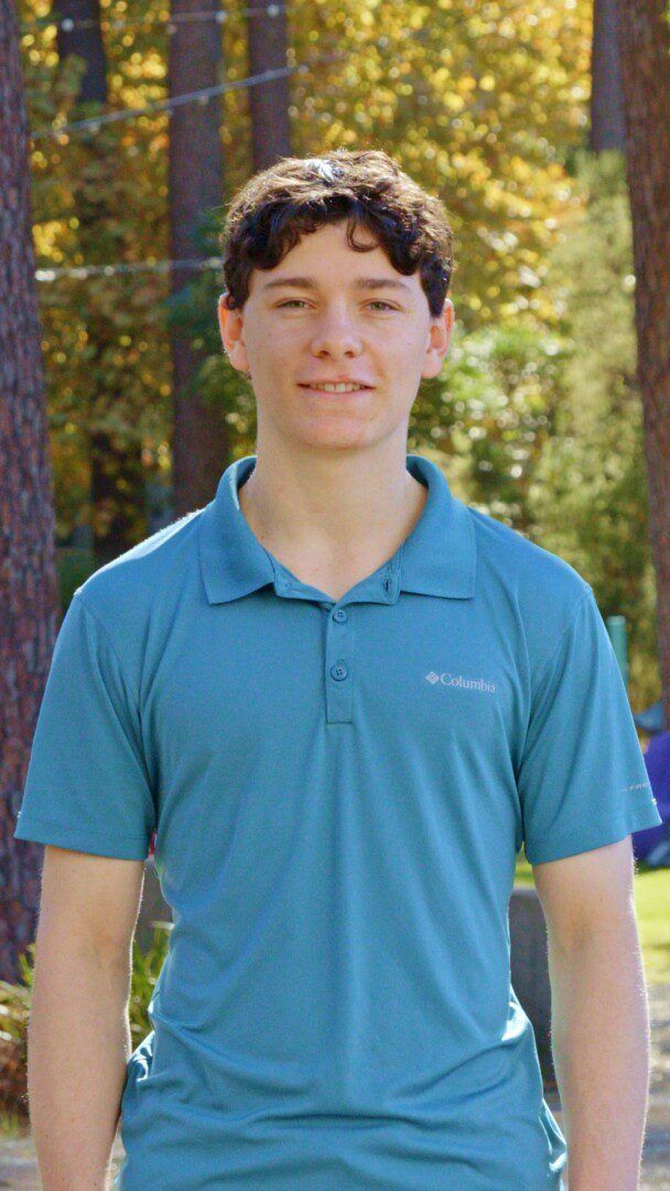 Portrait of a young man with curly brown hair, smiling, wearing a teal Columbia polo shirt, standing outdoors with trees in the background