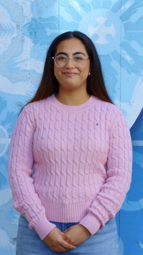 Portrait of a young woman with long dark hair and glasses, smiling, wearing a pink cable-knit sweater and jeans, standing in front of a blue mural.