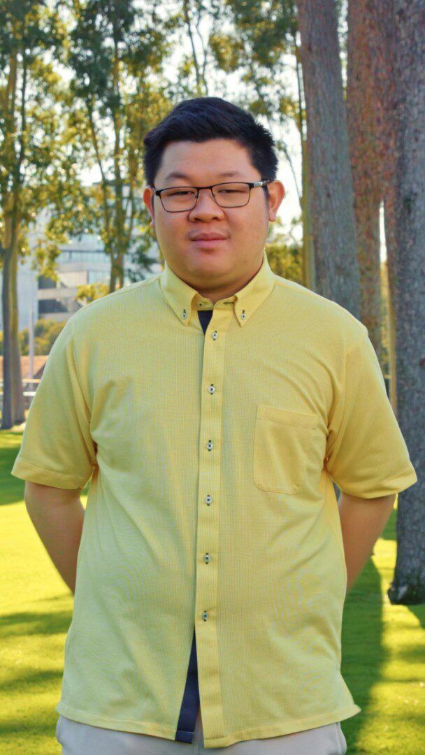 Portrait of a young man with glasses, wearing a pale yellow short-sleeve button-up shirt, standing outdoors on grass with trees in the background