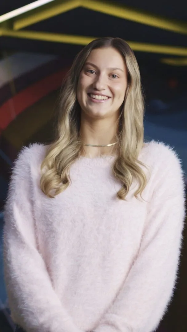 Portrait of a young woman with long blonde wavy hair, smiling, wearing a fluffy pale pink sweater, standing indoors in front of a colourful abstract mural.