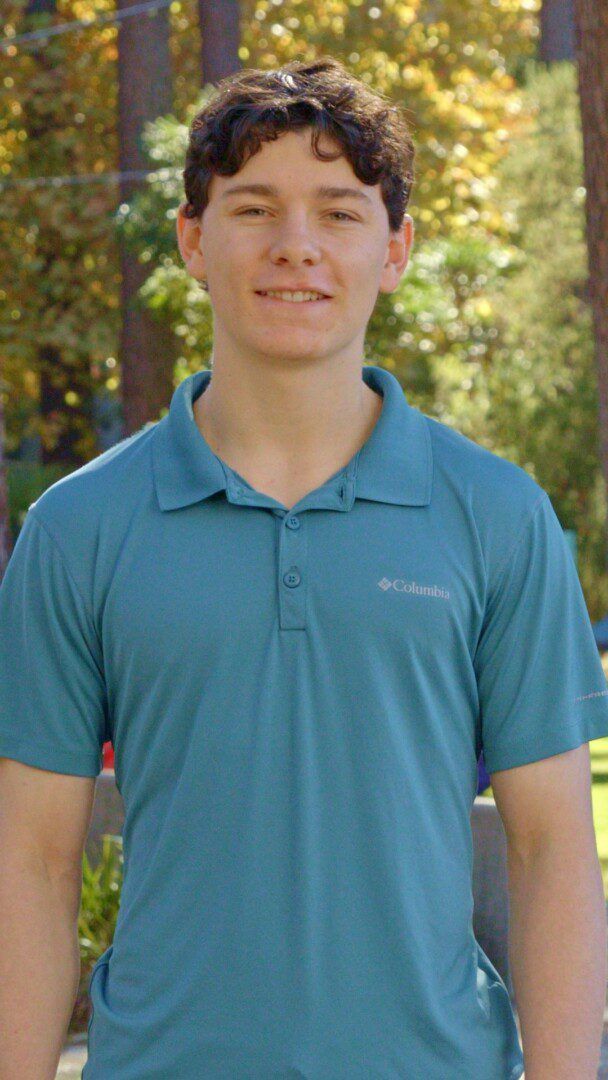 Portrait of a young man with curly brown hair, smiling, wearing a teal Columbia polo shirt, standing outdoors with trees in the background