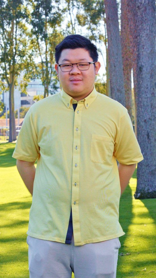 Portrait of a young man with glasses, wearing a pale yellow short-sleeve button-up shirt, standing outdoors on grass with trees in the background