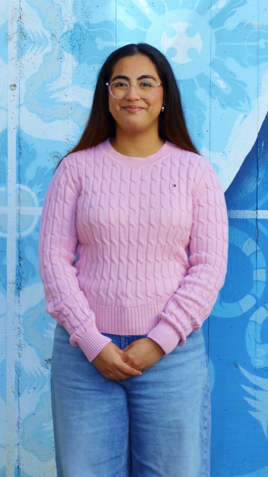Portrait of a young woman with long dark hair and glasses, smiling, wearing a pink cable-knit sweater and jeans, standing in front of a blue mural.