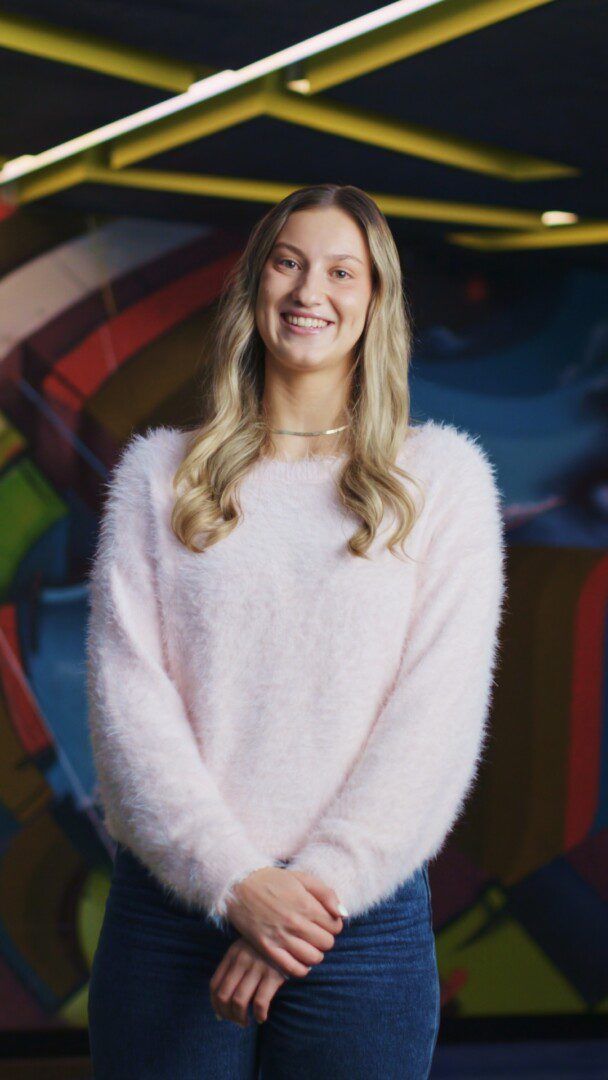 Portrait of a young woman with long blonde wavy hair, smiling, wearing a fluffy pale pink sweater, standing indoors in front of a colourful abstract mural.