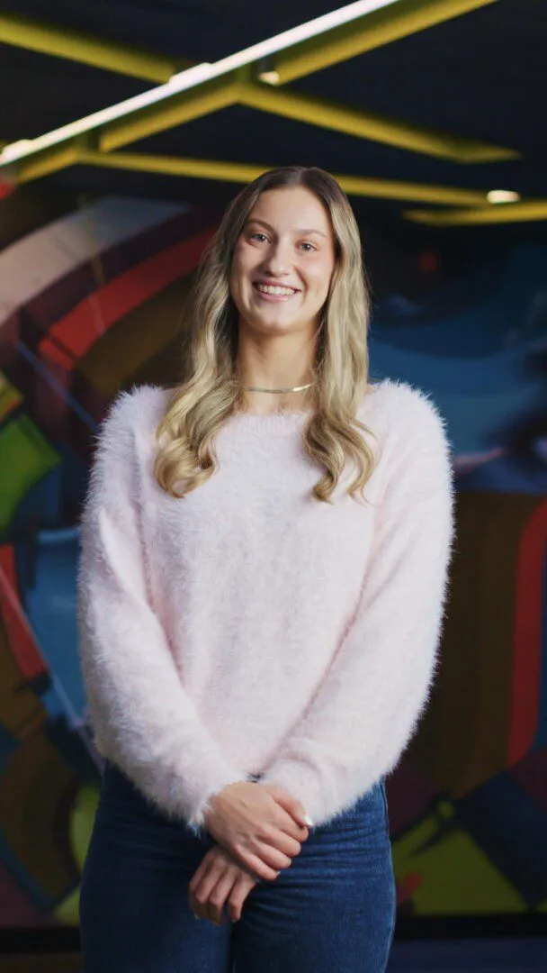 Portrait of a young woman with long blonde wavy hair, smiling, wearing a fluffy pale pink sweater, standing indoors in front of a colourful abstract mural.