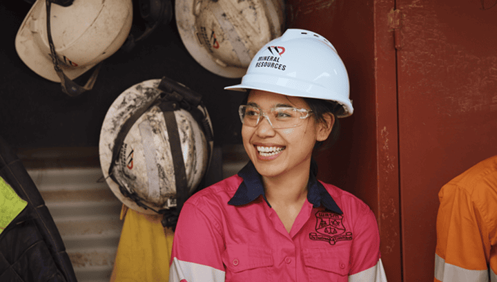 A student wearing a white hard hat and pink work shirt, smiling, standing in front of a row of hard hats on a rack