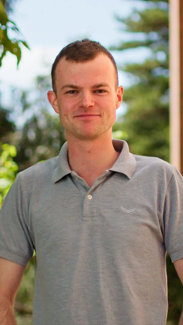 Hendryk smiling outdoors in a gray polo shirt, with green trees behind him.