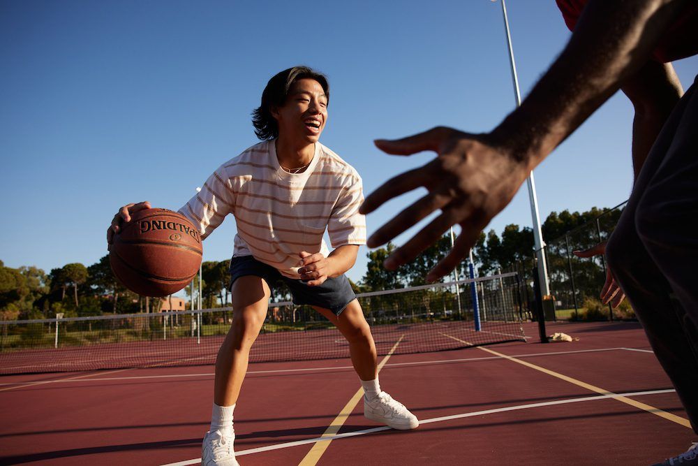 A young man laughs as he dribbles a basketball on an outdoor court, while a defender’s hand reaches towards the ball.